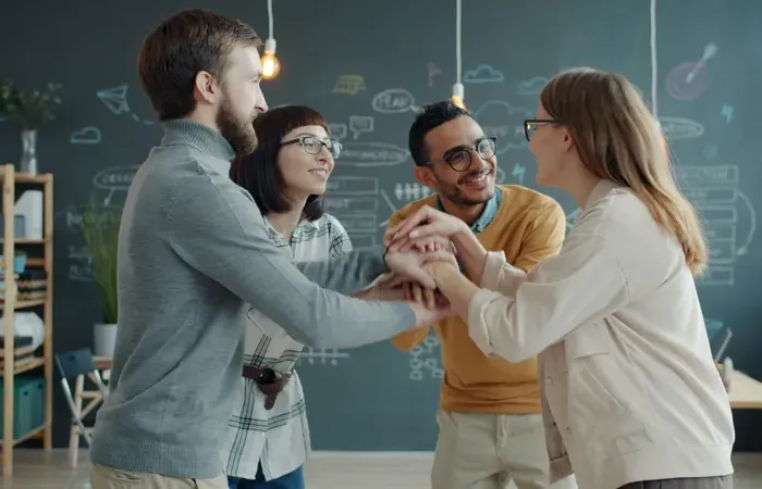 Diverse team of coworkers smiling and joining hands together in modern creative office with chalkboard wall